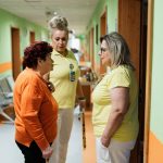 Two nurses interacting with a senior in a brightly colored care facility hallway.